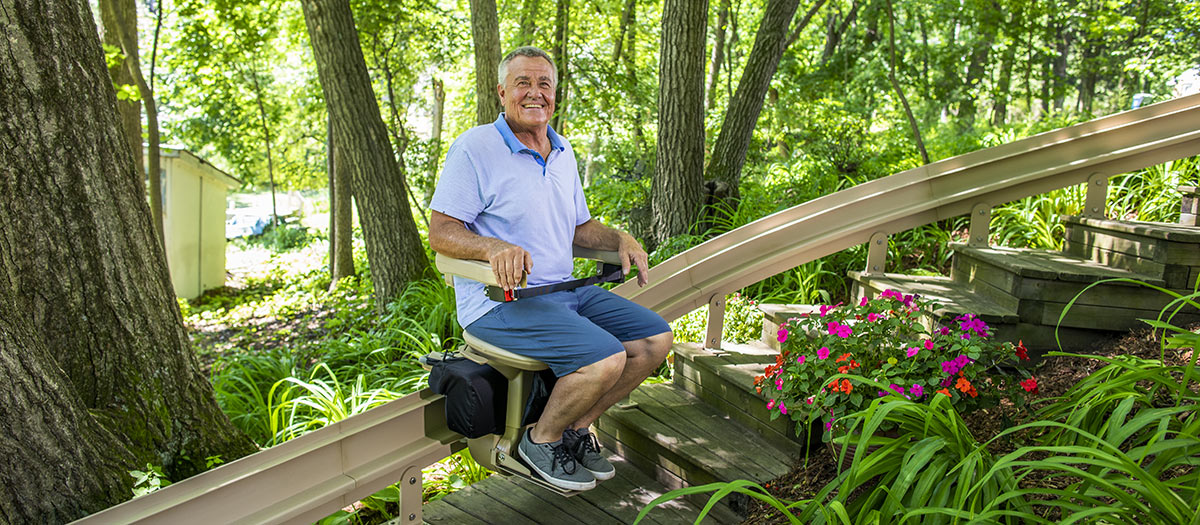 Man sitting on a Bruno Elite curved outdoor stairlift halfway up a wooden staircase near some trees