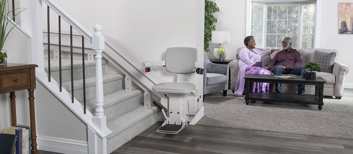 Bruno Elite Curved indoor stairlift parked at the bottom of a staircase with a husband and wife sitting on a couch