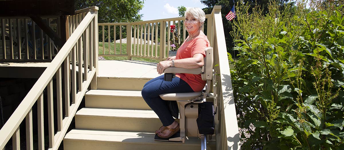 Woman sitting on a Bruno Elite straight outdoor stairlift halfway up the steps with an American flag in the background